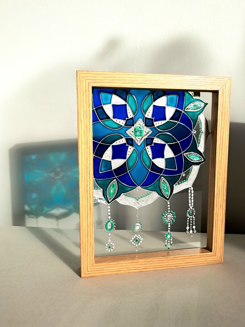 Op de afbeelding: Een ingelijst glas-in-lood kunstwerk met een geometrisch ontwerp in tinten blauw, turquoise en wit. Het kunstwerk bevat hangende juweelachtige versieringen in groen en zilver, geplaatst in een natuurlijke houten lijst.