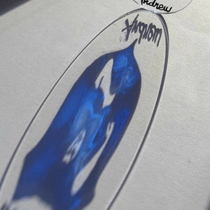 Op de afbeelding: Een blauwe en witte glazen ornament met een Schots vlagontwerp en de naam "Andrew" erop geschreven. De ornament hangt aan een zwart touwtje.