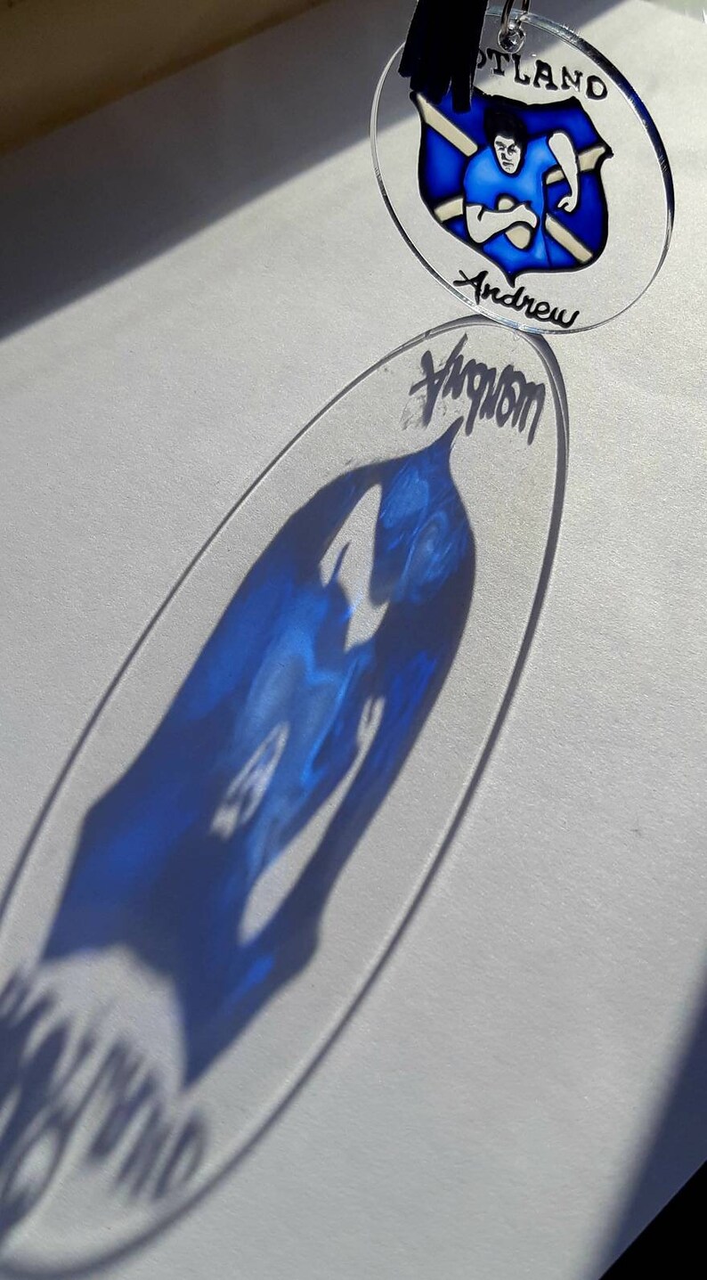 Op de afbeelding: Een blauwe en witte glazen ornament met een Schots vlagontwerp en de naam "Andrew" erop geschreven. De ornament hangt aan een zwart touwtje.