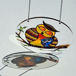 May include: A stained glass oval-shaped hanging ornament featuring a mother owl and baby owl. The mother owl is yellow, orange, and brown with intricate patterns. The baby owl is blue with large black eyes. The ornament is suspended by a silver chain.