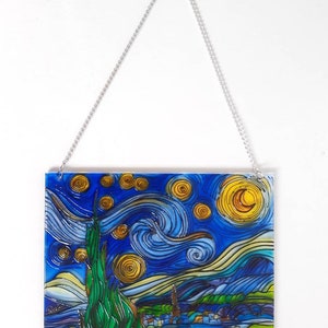 May include: A stained glass wall hanging featuring a recreation of Vincent van Gogh's "Starry Night" painting. The artwork depicts a swirling night sky with a bright yellow moon and stars, a dark green cypress tree, and a small village in the distance.