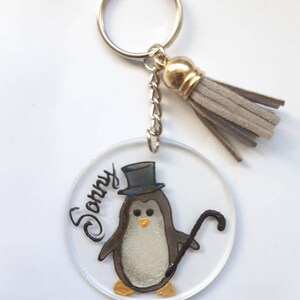 Op de afbeelding: Een ronde, transparante acryl sleutelhanger met een geschilderde pingu&iuml;n die een hoge hoed draagt en een stok vasthoudt. De pingu&iuml;n is grijs en wit met gouden accenten. De sleutelhanger heeft een grijze kwast en de naam "Sonny" staat op de sleutelhanger.