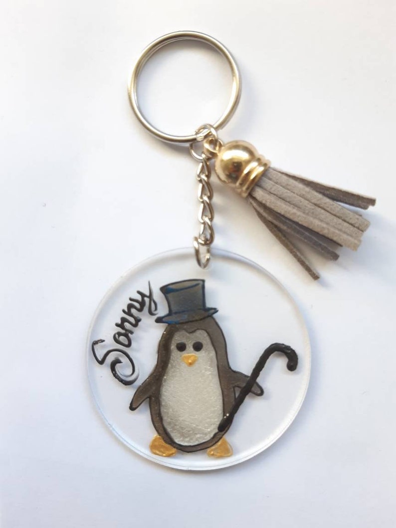 Op de afbeelding: Een ronde, transparante acryl sleutelhanger met een geschilderde pingu&iuml;n die een hoge hoed draagt en een stok vasthoudt. De pingu&iuml;n is grijs en wit met gouden accenten. De sleutelhanger heeft een grijze kwast en de naam "Sonny" staat op de sleutelhanger.