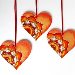 Op de afbeelding: Drie rode en oranje hartvormige ornamenten met een mandala-ontwerp erop geschilderd. De ornamenten hangen aan rode touwtjes.