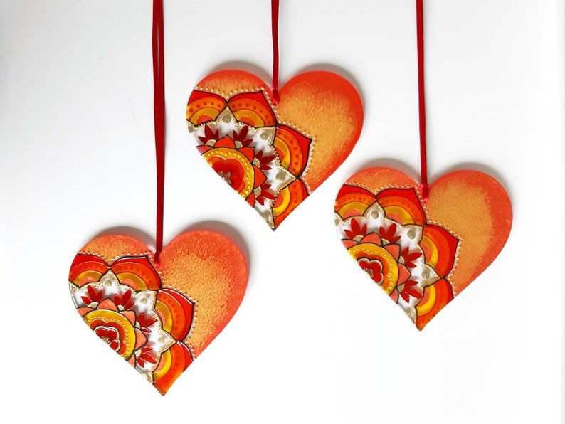 Op de afbeelding: Drie rode en oranje hartvormige ornamenten met een mandala-ontwerp erop geschilderd. De ornamenten hangen aan rode touwtjes.