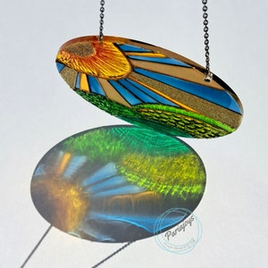 May include: A colorful oval-shaped suncatcher with a sun and green hills design. The suncatcher is hanging from a silver chain and casts a shadow on a white surface.