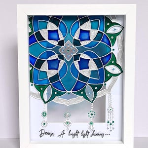 Op de afbeelding: Een ingelijst kunstwerk met een symmetrisch ontwerp in blauw, turquoise en groen. Het ontwerp bevat geometrische vormen, zilveren accenten en hangende versieringen. De tekst "Denize, A bright light shining..." staat onderaan.