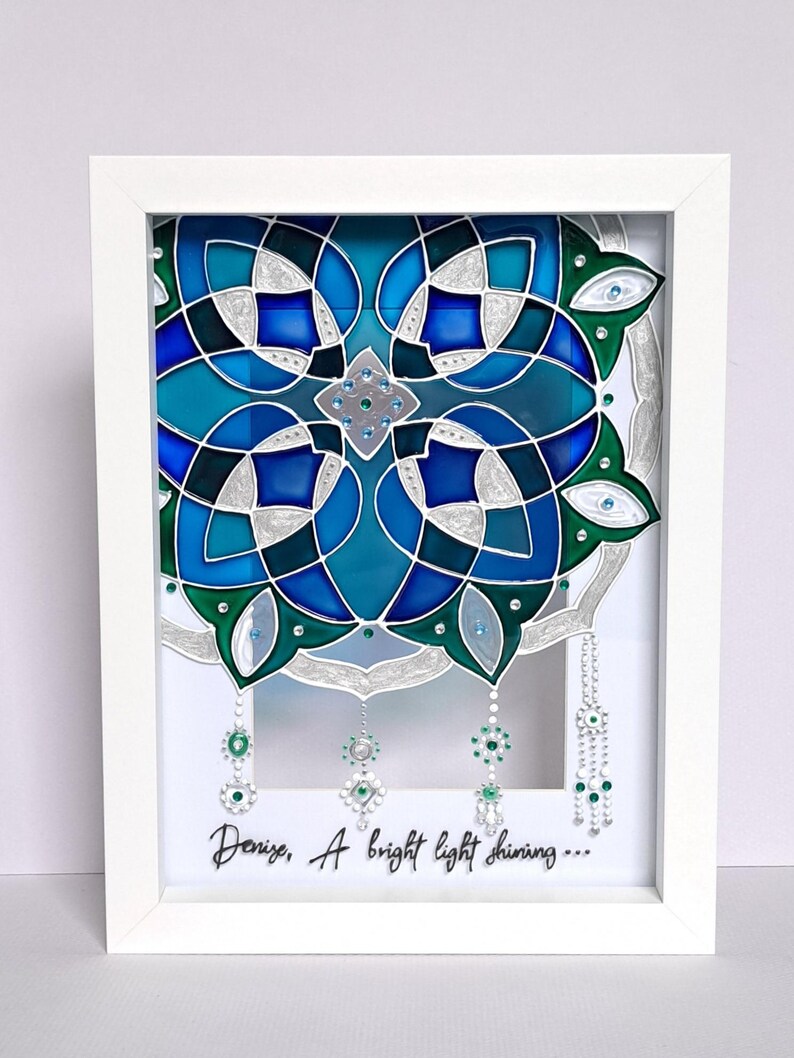 Op de afbeelding: Een ingelijst kunstwerk met een symmetrisch ontwerp in blauw, turquoise en groen. Het ontwerp bevat geometrische vormen, zilveren accenten en hangende versieringen. De tekst "Denize, A bright light shining..." staat onderaan.