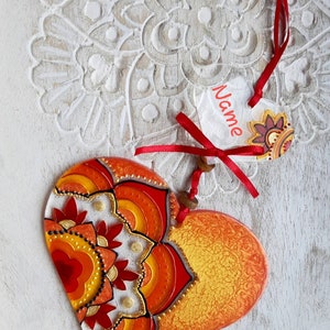 Op de afbeelding: Een hartvormige ornament in rood en oranje met een mandala-ontwerp. De ornament heeft een wit label eraan bevestigd met het woord "Naam" erop.