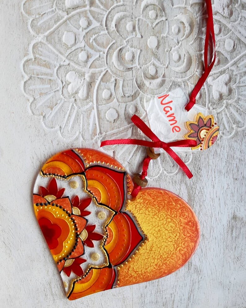 Op de afbeelding: Een hartvormige ornament in rood en oranje met een mandala-ontwerp. De ornament heeft een wit label eraan bevestigd met het woord "Naam" erop.