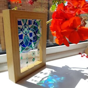 Op de afbeelding: Een ingelijst glas-in-lood kunstwerk met een geometrisch ontwerp in blauwe en groene tinten. De houten lijst is rechthoekig. Decoratieve hangende elementen zijn zichtbaar. Rode bloemen zijn op de achtergrond.