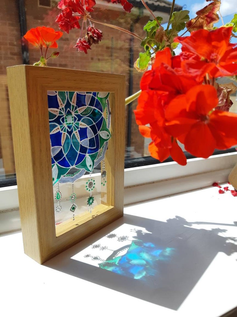 Op de afbeelding: Een ingelijst glas-in-lood kunstwerk met een geometrisch ontwerp in blauwe en groene tinten. De houten lijst is rechthoekig. Decoratieve hangende elementen zijn zichtbaar. Rode bloemen zijn op de achtergrond.