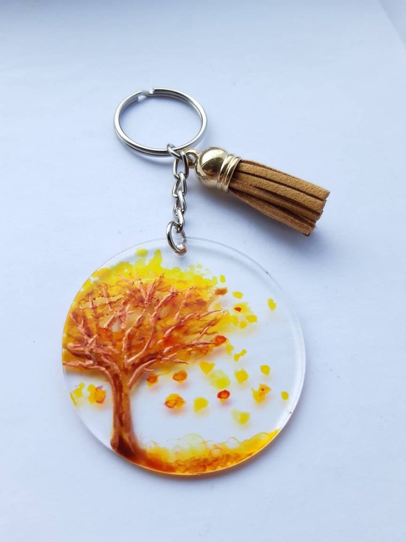Op de afbeelding: Een ronde sleutelhanger van hars met een geschilderd beeld van een boom met gele en oranje bladeren. De sleutelhanger heeft een gouden metalen ring en een bruine kwast.