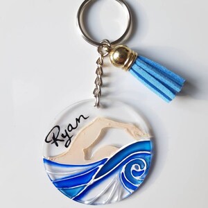 Op de afbeelding: Een ronde, transparante acryl sleutelhanger met een blauw en wit geschilderd ontwerp van een zwemmer in het water. De naam "Ryan" staat in het zwart op de sleutelhanger. De sleutelhanger heeft een zilveren sleutelring en een blauwe kwast.