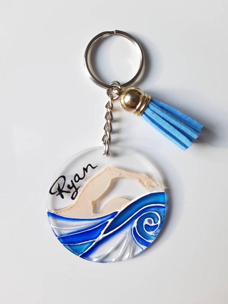 Op de afbeelding: Een ronde, transparante acryl sleutelhanger met een blauw en wit geschilderd ontwerp van een zwemmer in het water. De naam "Ryan" staat in het zwart op de sleutelhanger. De sleutelhanger heeft een zilveren sleutelring en een blauwe kwast.