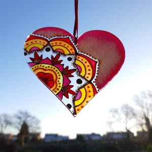 Op de afbeelding: Een roze glazen hartvormige ornament met een kleurrijk mandala-ontwerp. De mandala heeft rode, gele, oranje en roze kleuren. De ornament hangt aan een rood lint.