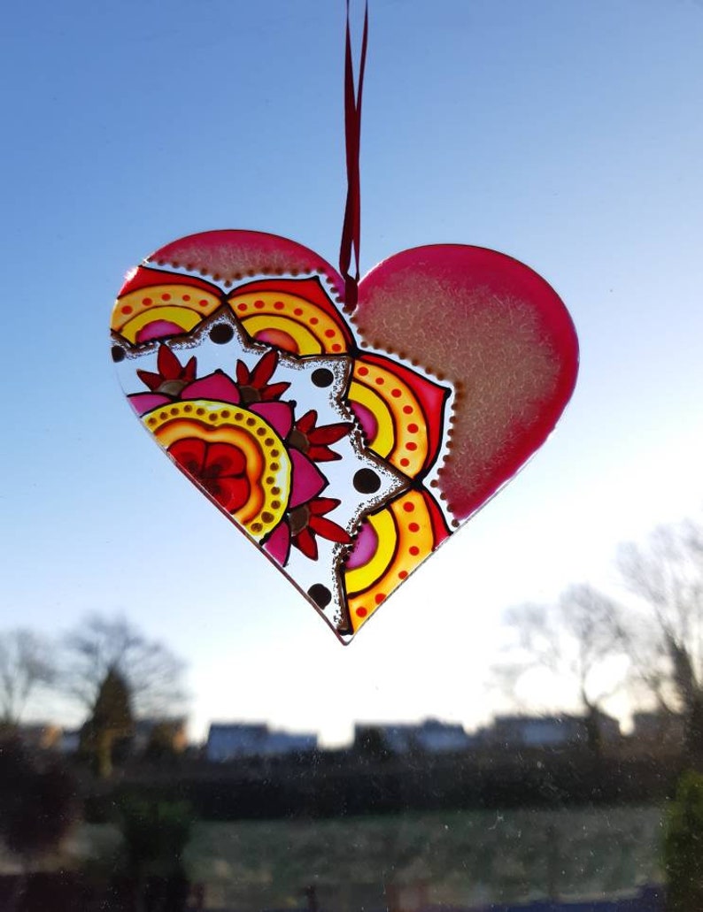 Op de afbeelding: Een roze glazen hartvormige ornament met een kleurrijk mandala-ontwerp. De mandala heeft rode, gele, oranje en roze kleuren. De ornament hangt aan een rood lint.