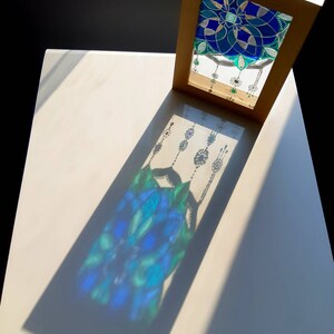Op de afbeelding: Een kunstwerk met een lijst met een glas-in-lood ontwerp. Het glas heeft een blauw en groen bloemenpatroon. Het zonlicht werpt een schaduw van het ontwerp op het witte oppervlak eronder. De lijst is lichtbruin van kleur.