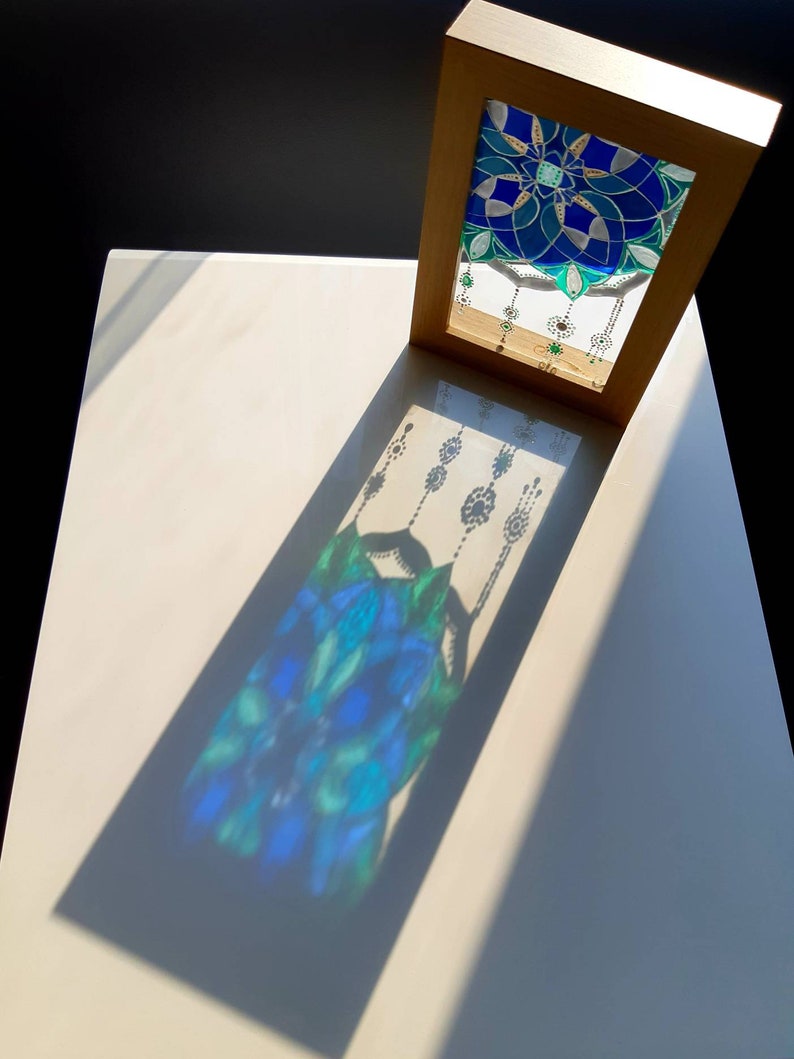 Op de afbeelding: Een kunstwerk met een lijst met een glas-in-lood ontwerp. Het glas heeft een blauw en groen bloemenpatroon. Het zonlicht werpt een schaduw van het ontwerp op het witte oppervlak eronder. De lijst is lichtbruin van kleur.