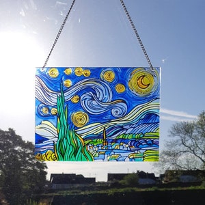 May include: A stained glass window hanging with a depiction of Vincent van Gogh's Starry Night. The artwork features a swirling blue sky with yellow stars and a crescent moon. The bottom of the image shows a small town with green trees and hills.