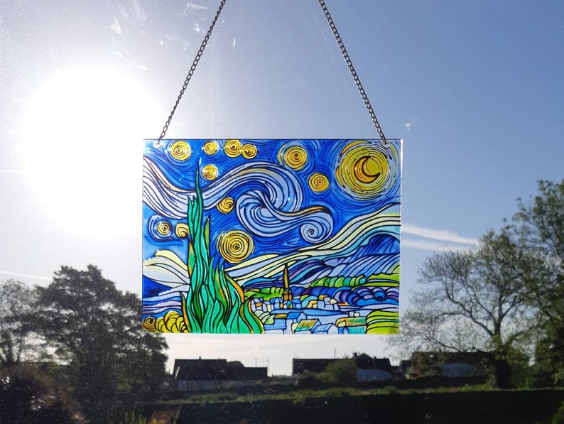 May include: A stained glass window hanging with a depiction of Vincent van Gogh's Starry Night. The artwork features a swirling blue sky with yellow stars and a crescent moon. The bottom of the image shows a small town with green trees and hills.