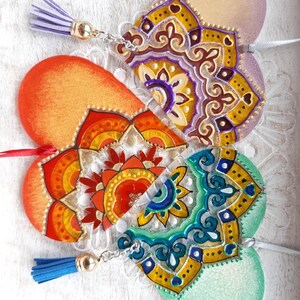 Op de afbeelding: Drie hartvormige houten ornamenten met geschilderde mandala-ontwerpen. De harten zijn verbonden door een zilveren ketting en hebben kwastjes. De kleuren van de mandala's zijn paars, oranje en groen.