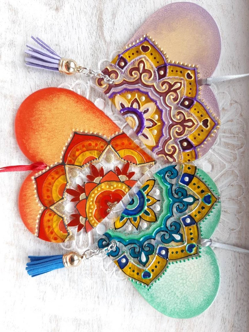 Op de afbeelding: Drie hartvormige houten ornamenten met geschilderde mandala-ontwerpen. De harten zijn verbonden door een zilveren ketting en hebben kwastjes. De kleuren van de mandala's zijn paars, oranje en groen.