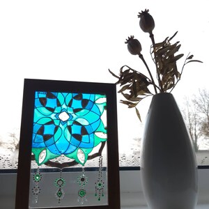 Op de afbeelding: Een ingelijst glas-in-loodpaneel met een geometrisch ontwerp in blauwe en groene tinten. Het paneel is versierd met hangende decoratieve elementen. Een witte vaas met gedroogde bloemen staat ernaast, wat een decoratieve opstelling cre&euml;ert.