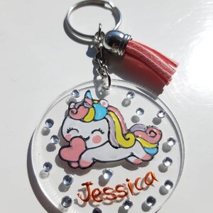 Op de afbeelding: Een transparante acryl sleutelhanger met een eenhoorn ontwerp en de naam "Jessica" in oranje glitter. De sleutelhanger heeft een roze kwast en een zilveren sleutelring.
