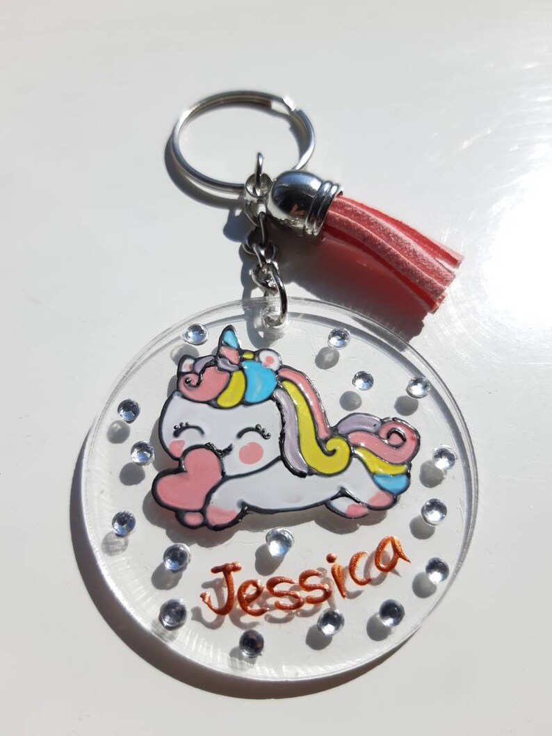 Op de afbeelding: Een transparante acryl sleutelhanger met een eenhoorn ontwerp en de naam "Jessica" in oranje glitter. De sleutelhanger heeft een roze kwast en een zilveren sleutelring.
