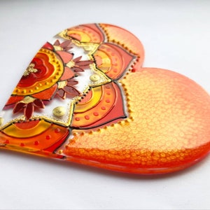 Op de afbeelding: Een hartvormige harsversiering met een kleurrijk mandala-ontwerp in tinten rood, oranje en geel. De mandala is omgeven door een gouden glitterrand.