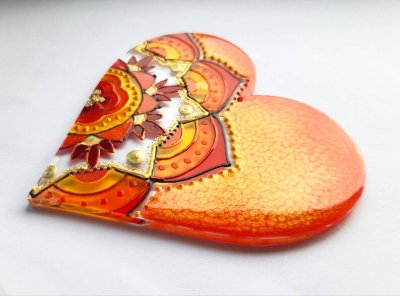 Op de afbeelding: Een hartvormige harsversiering met een kleurrijk mandala-ontwerp in tinten rood, oranje en geel. De mandala is omgeven door een gouden glitterrand.