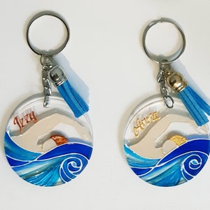 Op de afbeelding: Twee ronde, blauwe en witte sleutelhangers van hars met een zwemmer silhouet en een gouden zon. De sleutelhangers hebben een zilveren ring en een blauwe kwast. De sleutelhangers zijn gepersonaliseerd met de namen "Izzy" en "Olivia".