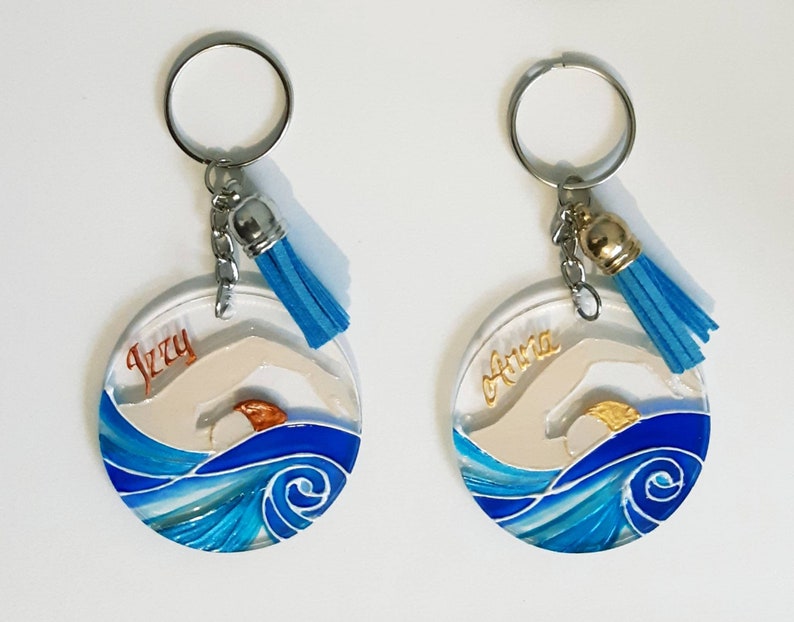 Op de afbeelding: Twee ronde, blauwe en witte sleutelhangers van hars met een zwemmer silhouet en een gouden zon. De sleutelhangers hebben een zilveren ring en een blauwe kwast. De sleutelhangers zijn gepersonaliseerd met de namen "Izzy" en "Olivia".