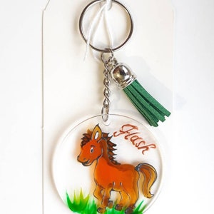 Op de afbeelding: Een ronde, transparante acryl sleutelhanger met een handgeschilderd cartoonpaard in oranje en bruin. Het paard staat op groen gras. De sleutelhanger heeft een zilveren ring en een groene kwast. Het woord "Flash" staat op de sleutelhanger.