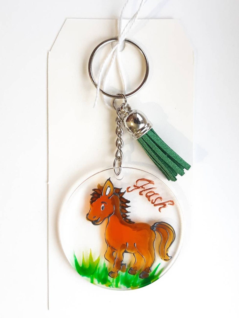 Op de afbeelding: Een ronde, transparante acryl sleutelhanger met een handgeschilderd cartoonpaard in oranje en bruin. Het paard staat op groen gras. De sleutelhanger heeft een zilveren ring en een groene kwast. Het woord "Flash" staat op de sleutelhanger.