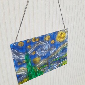 May include: A glass wall hanging featuring a painting of Vincent van Gogh's Starry Night. The painting is done in shades of blue, yellow, and green, with a swirling sky and a bright yellow moon.