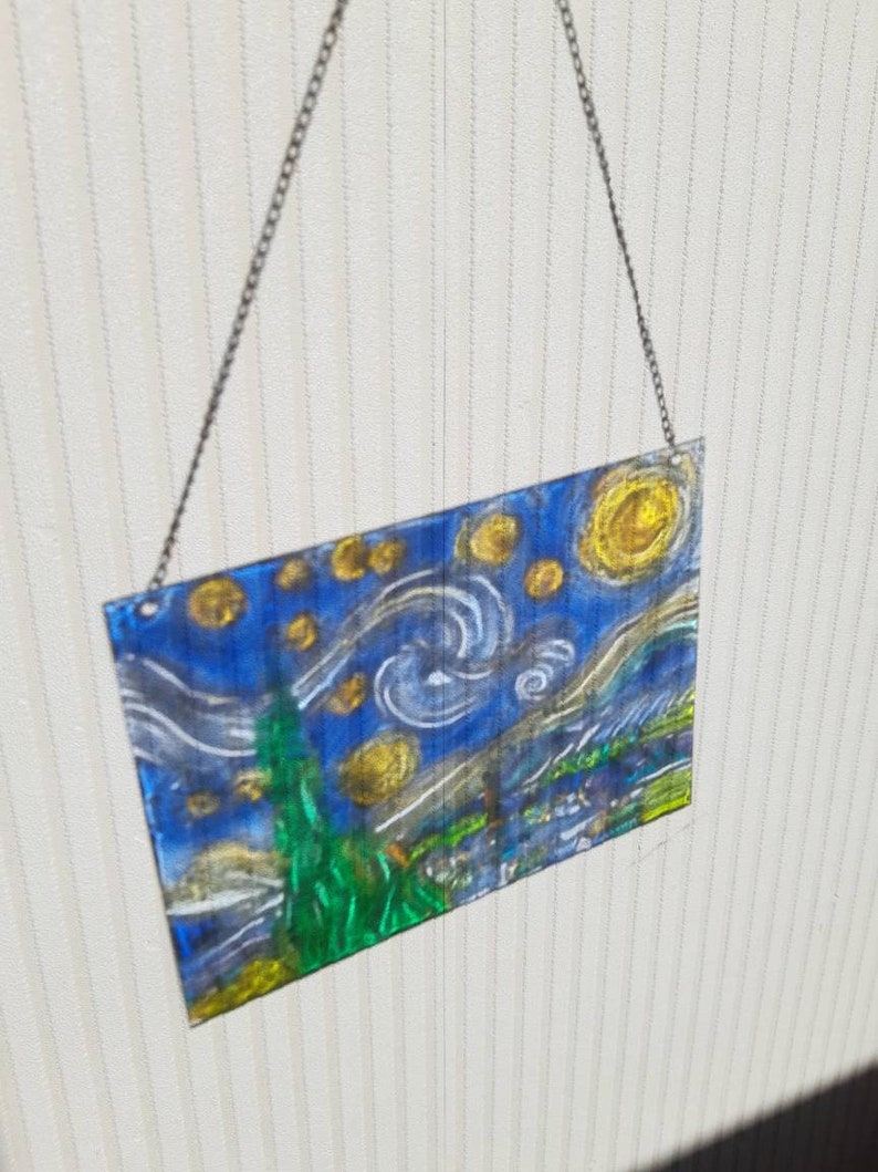 May include: A glass wall hanging featuring a painting of Vincent van Gogh's Starry Night. The painting is done in shades of blue, yellow, and green, with a swirling sky and a bright yellow moon.