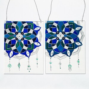 Op de afbeelding: Twee vierkante glazen panelen met ingewikkelde, symmetrische ontwerpen in blauw, groen en zilver. Elk paneel heeft een zilveren ketting om op te hangen en bungelende kralenaccenten. Het kunstwerk staat op een witte achtergrond.