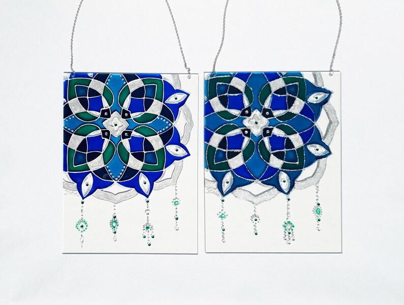 Op de afbeelding: Twee vierkante glazen panelen met ingewikkelde, symmetrische ontwerpen in blauw, groen en zilver. Elk paneel heeft een zilveren ketting om op te hangen en bungelende kralenaccenten. Het kunstwerk staat op een witte achtergrond.