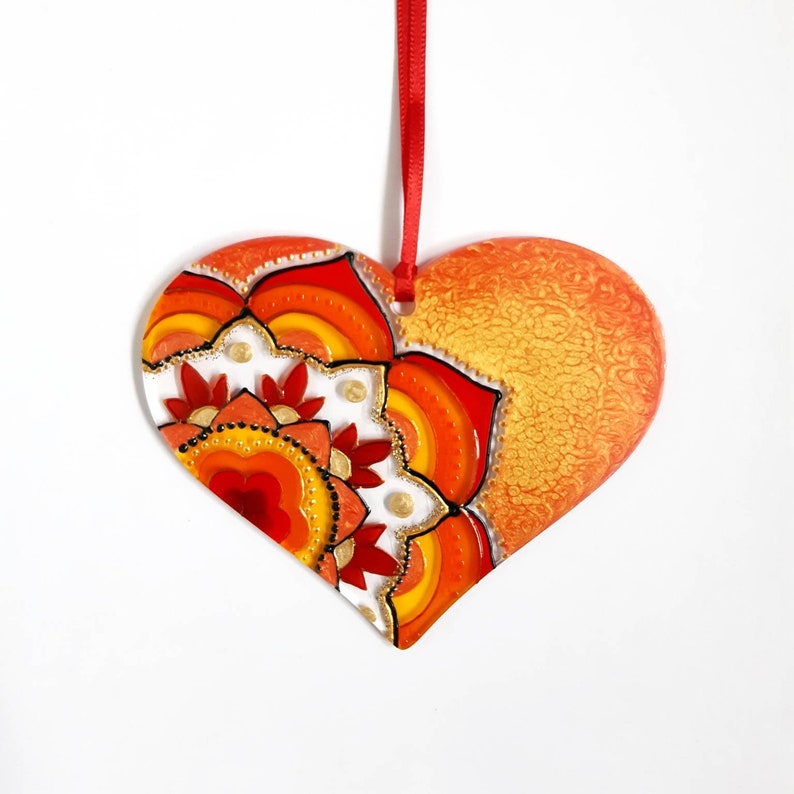 Op de afbeelding: Een hartvormige glazen ornament met een rood, oranje en geel mandala-ontwerp. De ornament heeft een gouden metallic afwerking en hangt aan een rood lint.
