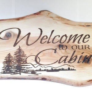 May include: Wooden sign with the text "Welcome to our Cabin" and a woodburned image of a forest scene with trees, mountains, and a moon.