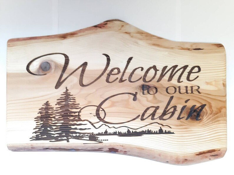 May include: Wooden sign with the text "Welcome to our Cabin" and a woodburned image of a forest scene with trees, mountains, and a moon.