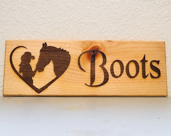 May include: Wooden sign with a silhouette of a girl riding a horse inside a heart shape. The text "Boots" is engraved on the right side of the sign.