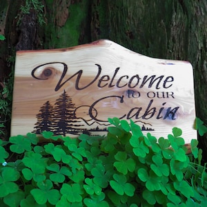 May include: A wooden sign with the text "Welcome to our Cabin" burned into the surface. The sign is in front of a green leafy background.