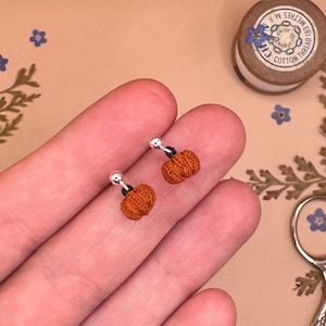 May include: A pair of orange pumpkin-shaped stud earrings. Each earring has a small, textured pumpkin design with a black stem and a silver post. The earrings are shown against a neutral background.