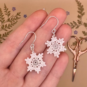 May include: Handmade white crochet snowflake earrings with silver-coloured hooks. The earrings have a delicate, lacy design. Each snowflake is centered with a small, silver-coloured bead. The earrings are displayed against a neutral background.