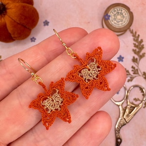 Könnte beinhalten: Ein Paar orangefarbene gehäkelte Blattohrringe mit goldfarbenen Akzenten. Die Ohrringe haben ein detailliertes Blattdesign mit einem goldenen Mittelteil und goldfarbenen Ohrhaken. Die Ohrringe sind ein Accessoire zum Thema Herbst.