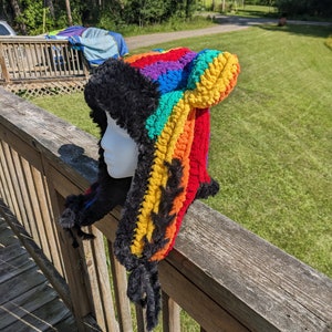 May include: A rainbow striped crocheted hat with black faux fur trim and ear flaps. The hat is on a wooden railing.