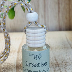 May include: A car air freshener with a clear glass bottle and a wooden cap. The label reads "Sunset Isle Car Diffuser | 0.30 oz." A braided cord with gold accents is attached for hanging. The background is a wooden surface.